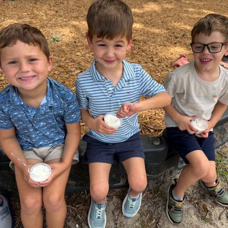 Three young boys seated on a bench in an outdoor setting. They hold small containers in their hands.
