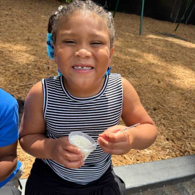 A young child wearing a striped tank top smiles and holds a container of yogurt in an outdoor playground.