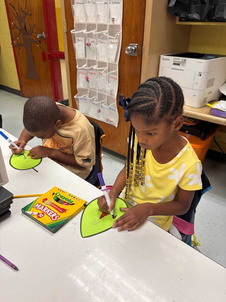 Children seated at a table. One holds a green paper heart while the other holds a crayon.