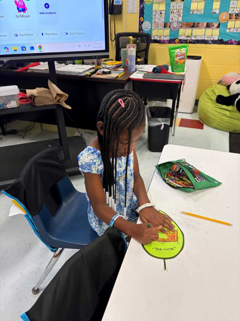 Child sitting at a desk in a classroom, working on a green circular project with pencils.
