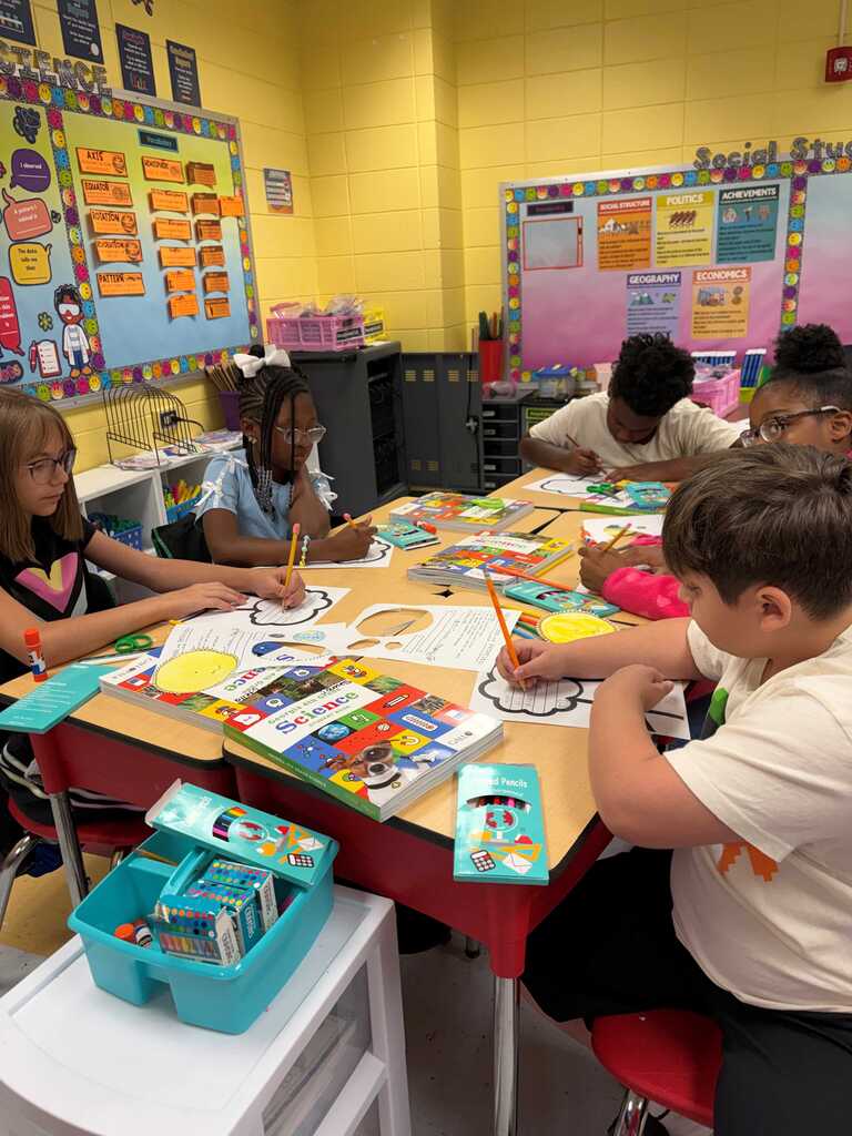 A group of children sitting at a table doing art projects, with a yellow wall and bulletin boards in the background.
