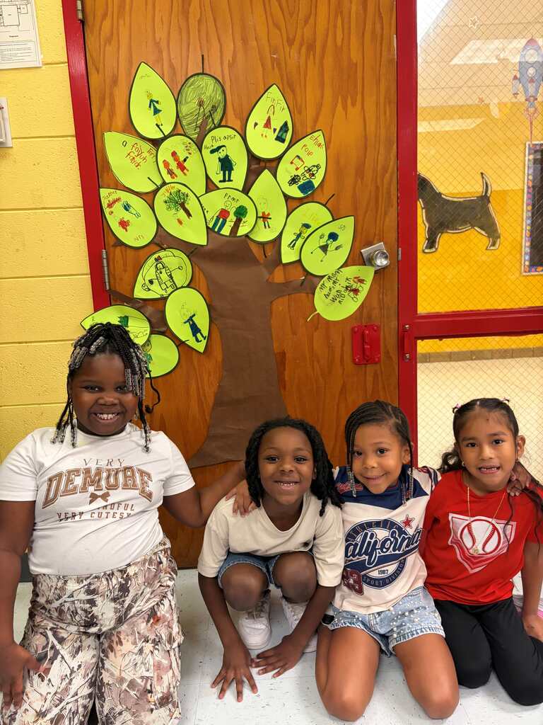 Four girls in a room, smiling. Behind them is a door with a tree illustration. The door is yellow and has a red border.