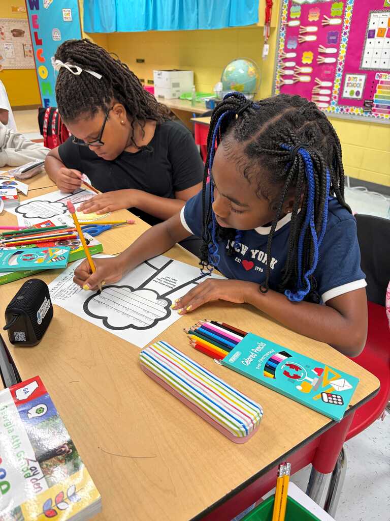 Two young girls seated at a desk, one coloring a picture with crayons, the other holding a pencil.