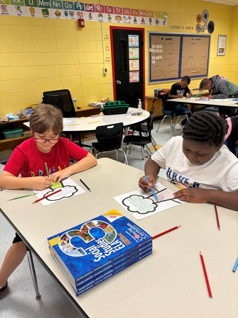 Two children sit at a table in a classroom, drawing on paper. A book titled "60 Years" sits on the table.