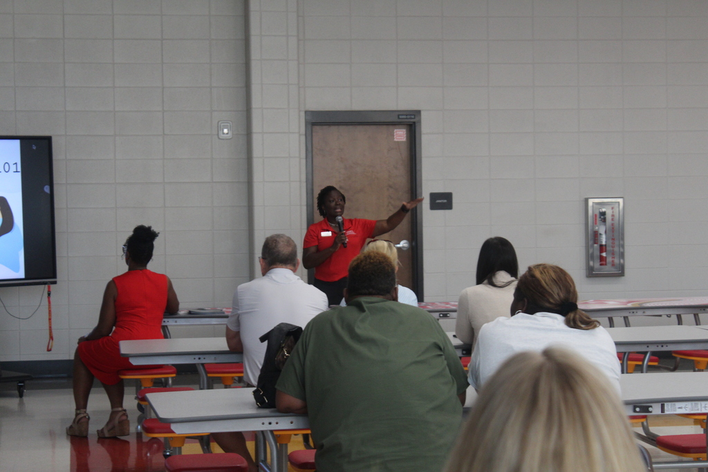 A group of people seated in chairs face a person standing in front of a screen giving a presentation.
