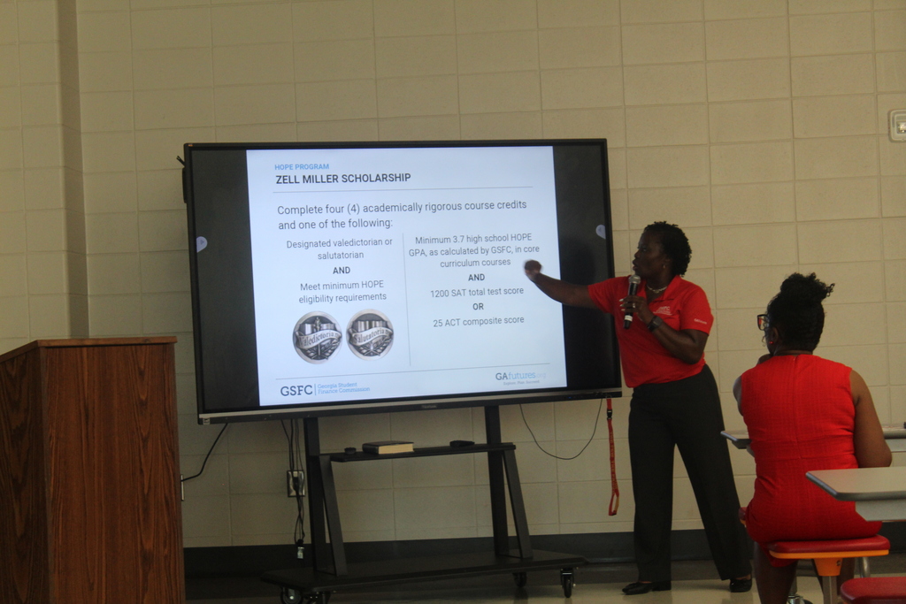 Woman in red shirt stands at podium, gesturing to a screen displaying a scholarship presentation. Audience seated.