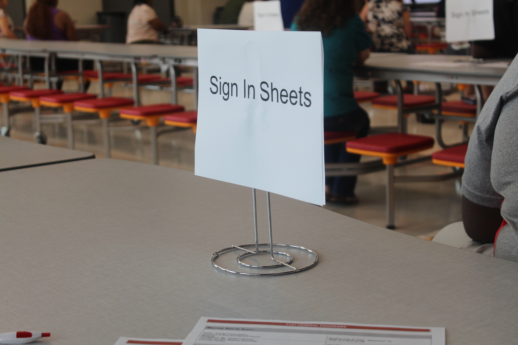 Sign on white paper reads "Sign In Sheets" on a stand. Tables and chairs are visible in the background.