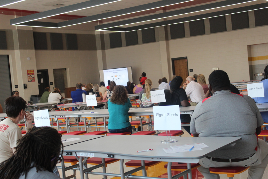 Room with tables and chairs, several people seated. A projector screen in the front displays a presentation. Each person has a small whiteboard for notes.