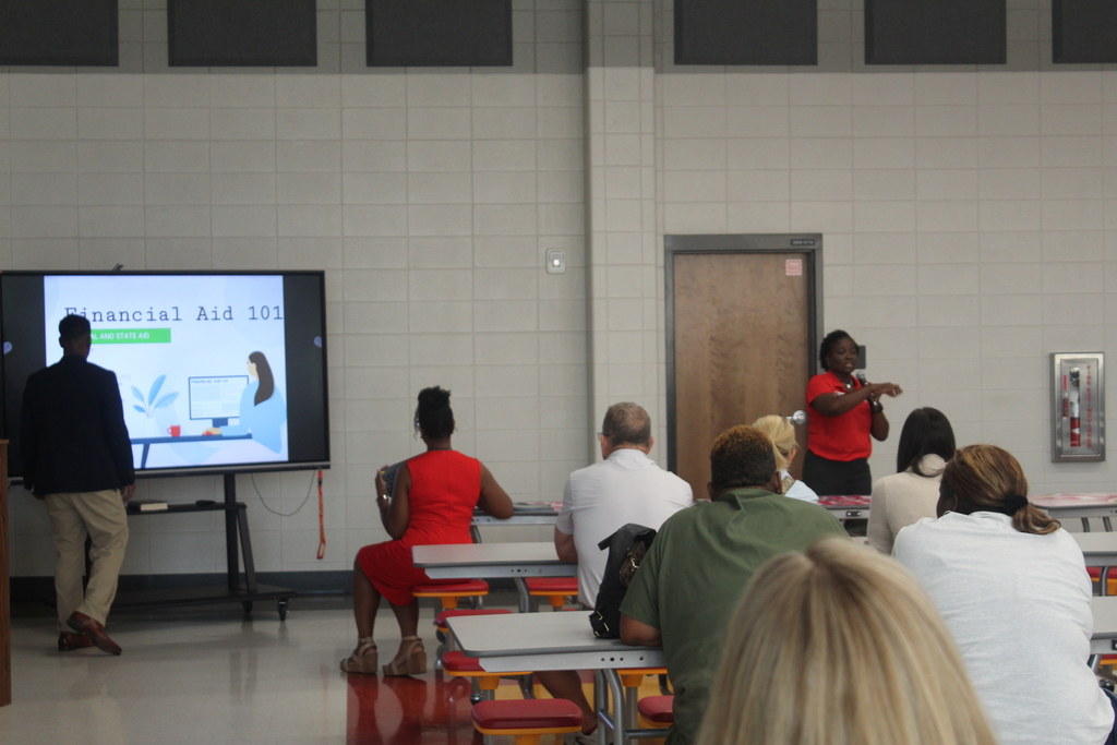 Room with a large screen; a woman speaks to a seated group, including a woman in red.