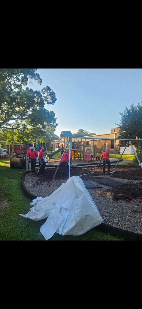Workers in red uniforms work in a park. A white tarp lies on the ground. Trees and a building are in the background.