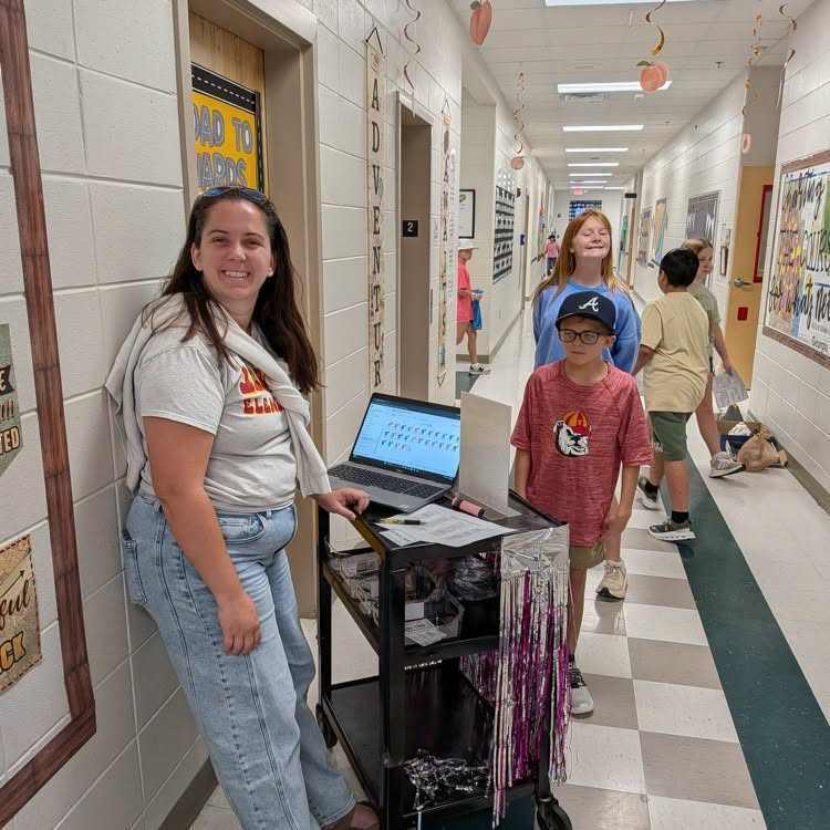 A woman with a laptop on a cart stands in a hallway. Two kids walk past, one wearing a cap. Bright walls and checkered floors.