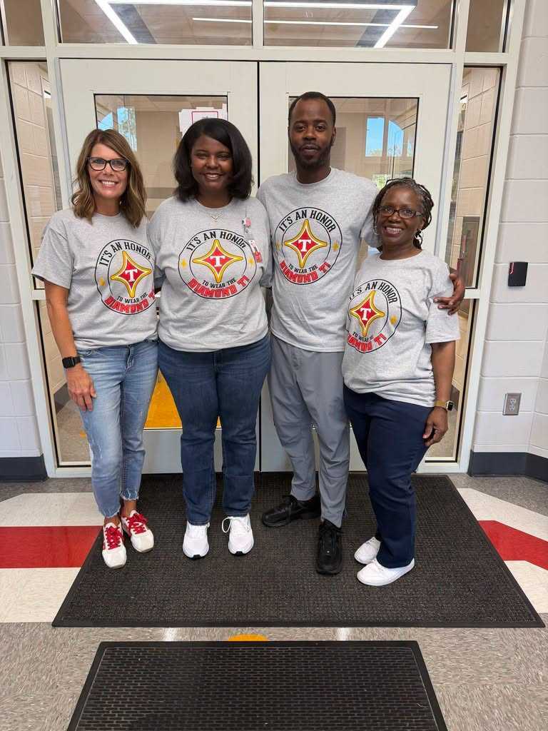 Four people stand on a black mat in front of a glass door. They wear gray shirts with a red logo. The floor is white and red.