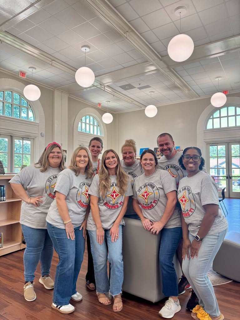 A group of people in matching gray t-shirts stands together in a room with wooden floors and a white ceiling.