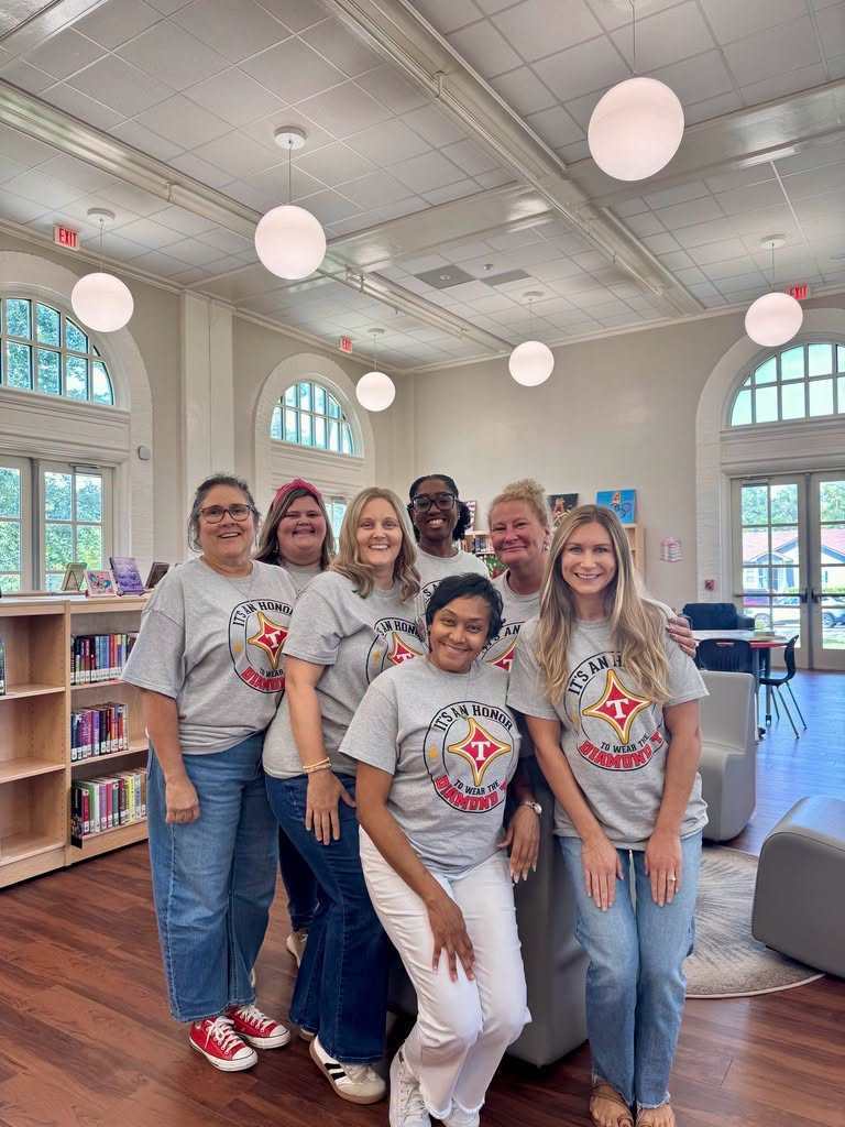 Group of adults, wearing matching shirts, standing in a room with wooden floors. Behind them, chairs, shelves, and windows.