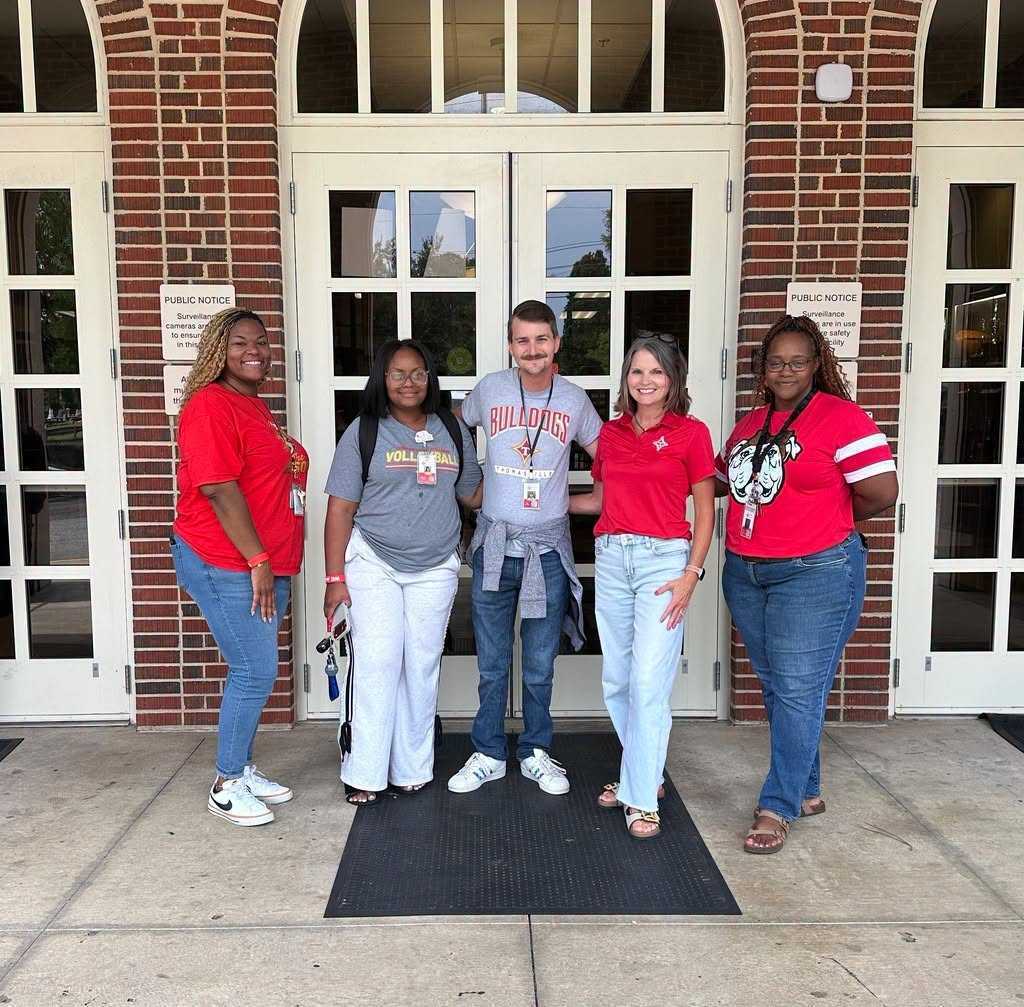 Five people stand outside a brick building. They wear casual clothing, with two wearing red shirts.