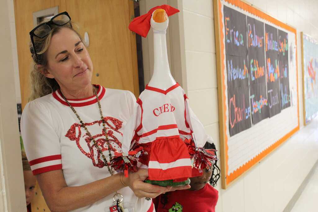 A woman wearing a white shirt with a red logo holds a red and white stuffed goose in a hallway.