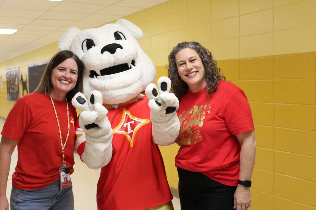 Three people in red shirts, including a mascot, stand in a hallway. The mascot wears a white costume.