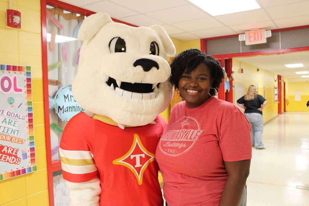 A woman and a mascot pose for a photo in a school hallway. Behind them is a classroom door and a bulletin board.