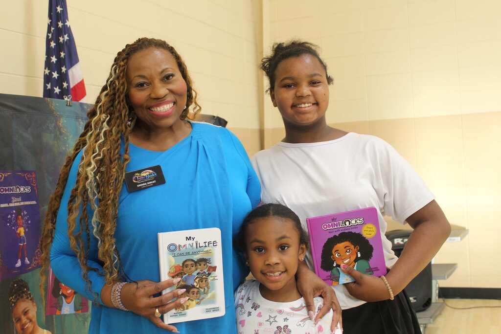 Three people stand together, smiling. A woman holds a book, and a young girl holds a book. Behind them, a flag is visible.