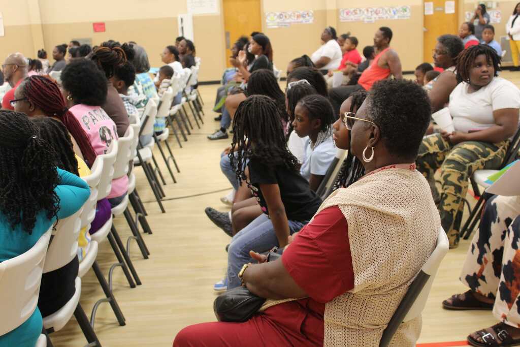 People seated in chairs at an event in a hall, some facing forward while others converse.