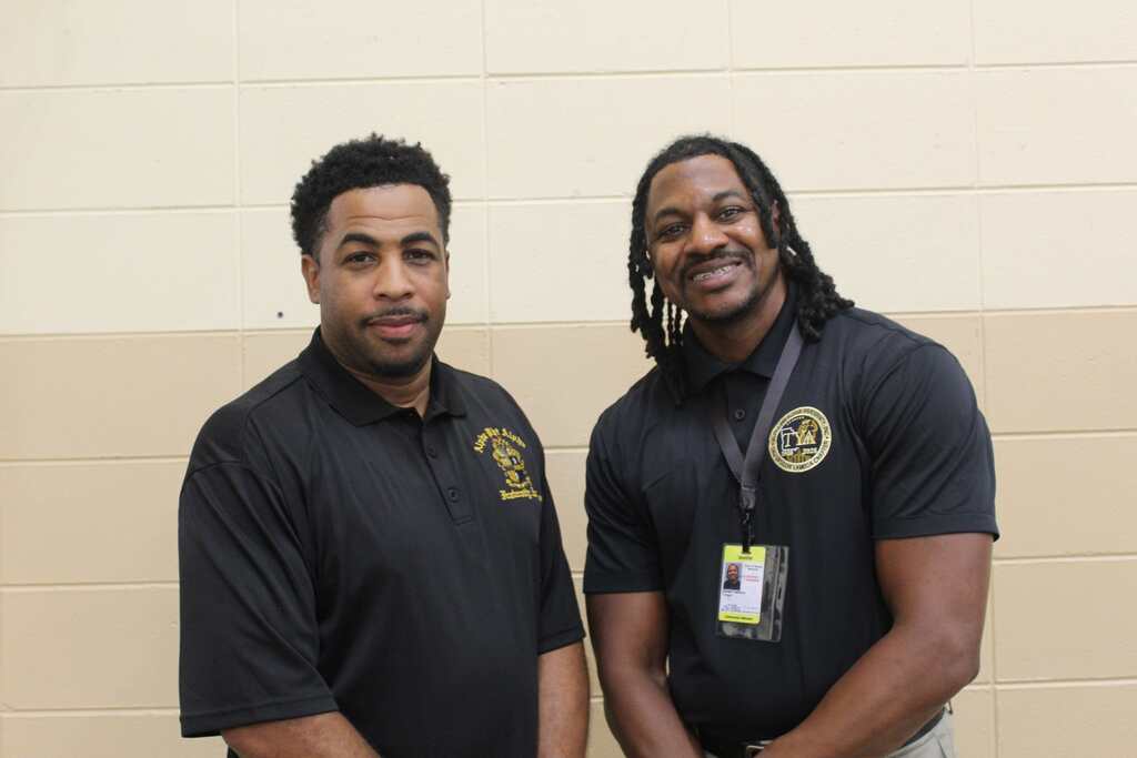 Two men in black polo shirts, one with a yellow logo on the left chest, pose for a picture.