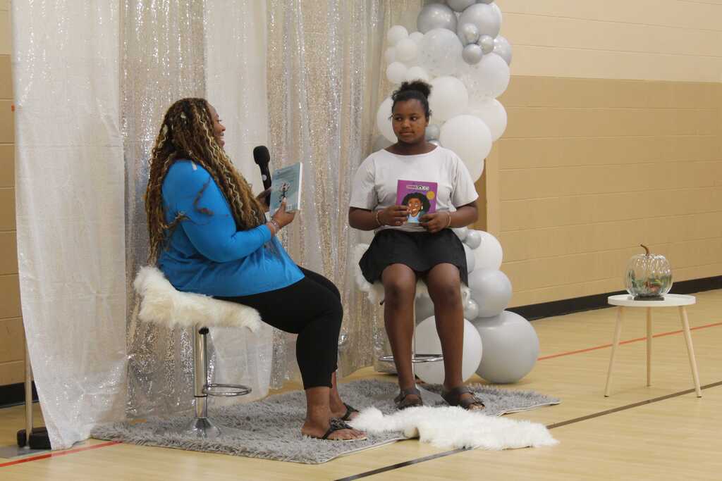 Two people seated, one holding a book, the other with a microphone, both facing each other. Behind them, a backdrop of white fabric and balloons.