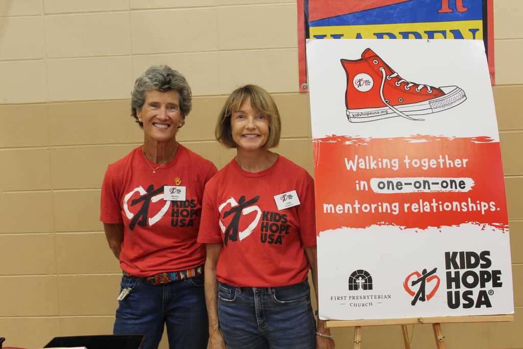 Two women in red shirts stand next to a sign with an image of a shoe. The sign reads, "Walking together in one-on-one mentoring relationships."