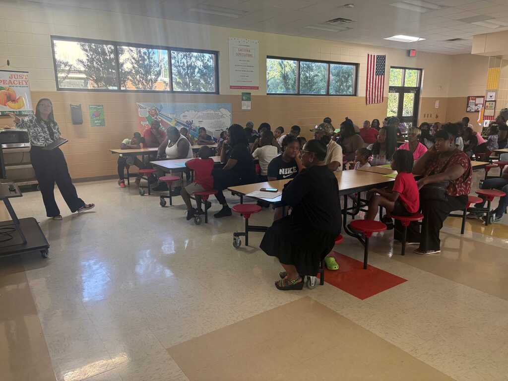 A school dining area with tables and chairs. A person stands near a table while others sit.