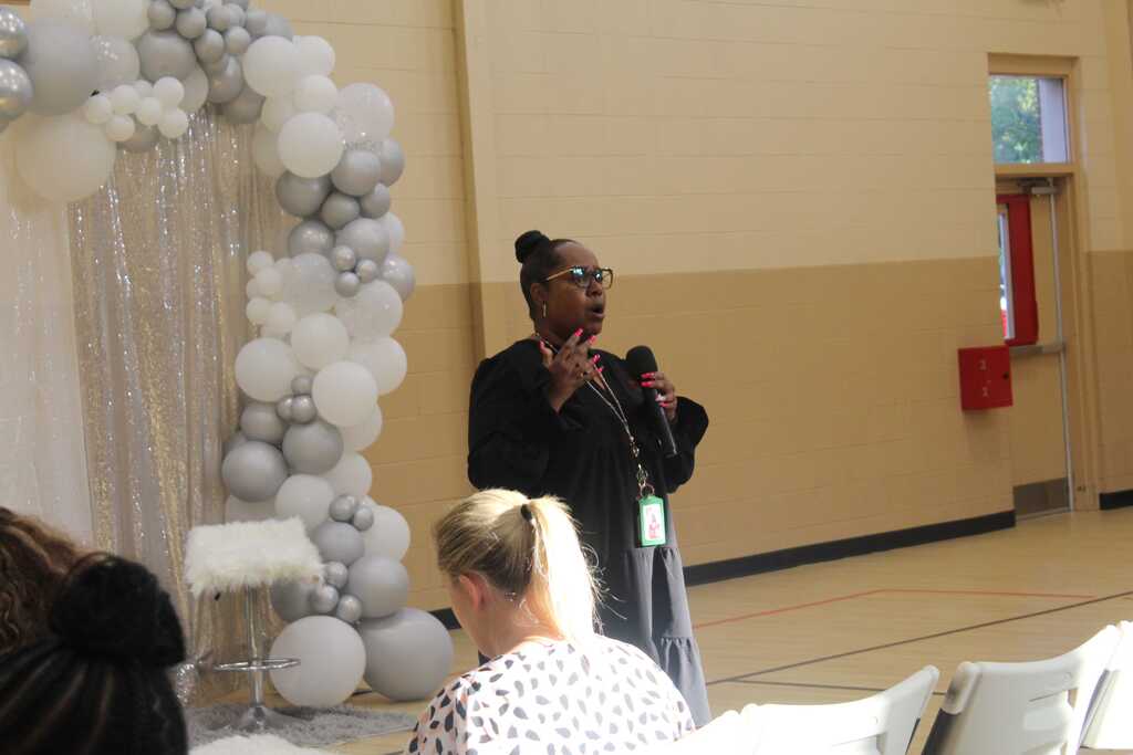 A person speaks into a microphone, standing before a large balloon arch, as two women look on.