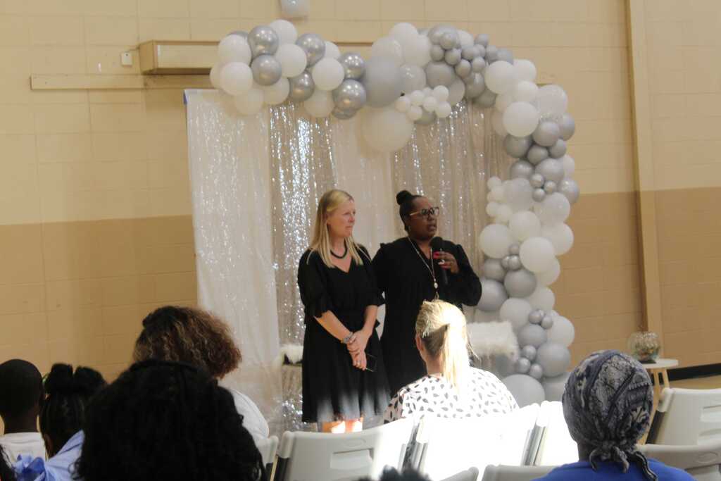 Two women stand behind a white balloon arch, addressing a group of seated attendees. The arch is composed of white balloons.