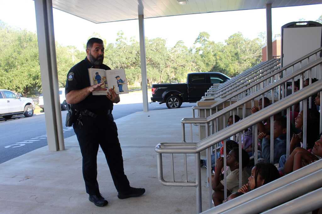 A uniformed officer holds a book, standing before a group of children seated on stairs outdoors.