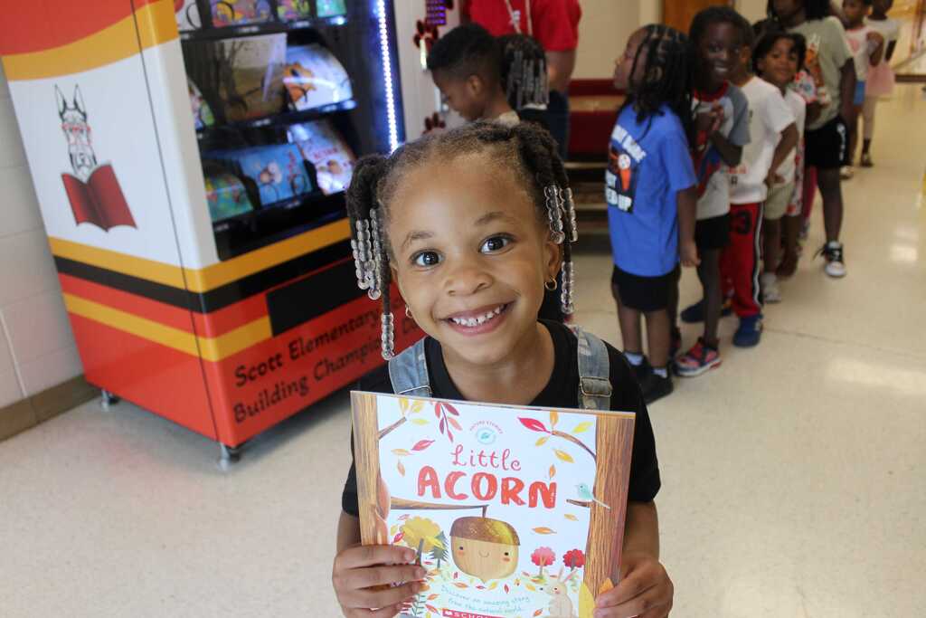 A young child holding a book titled "Little Acorn" stands in a school hallway, with children lined up behind.