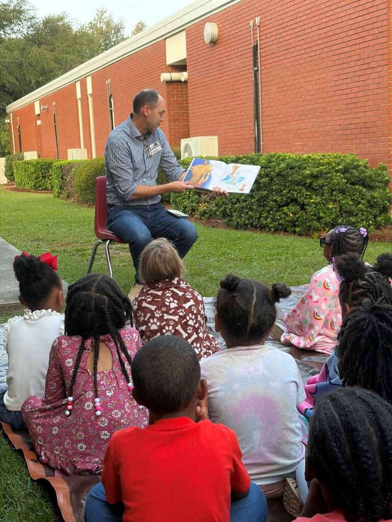Man sitting on a chair reading to a group of children seated in a semi-circle in front of him.