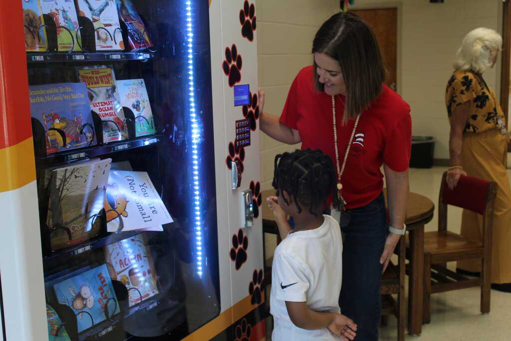 A woman with a red shirt assists a young child in choosing a book from a brightly lit vending machine.