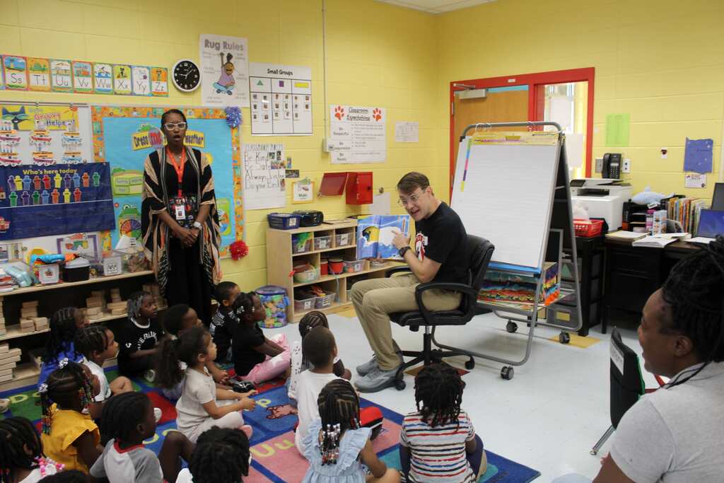 An indoor classroom with yellow walls and shelves full of books. A man plays guitar while seated, with many children in front.