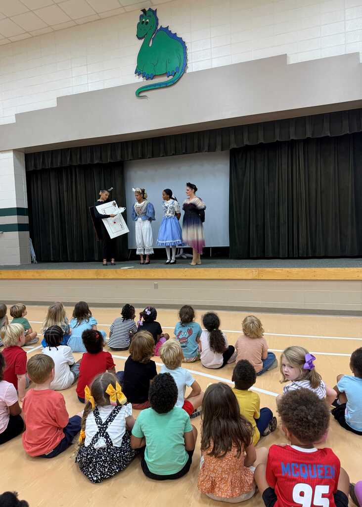 Several children sit in a circle, watching three people on a stage. One person holds a book.