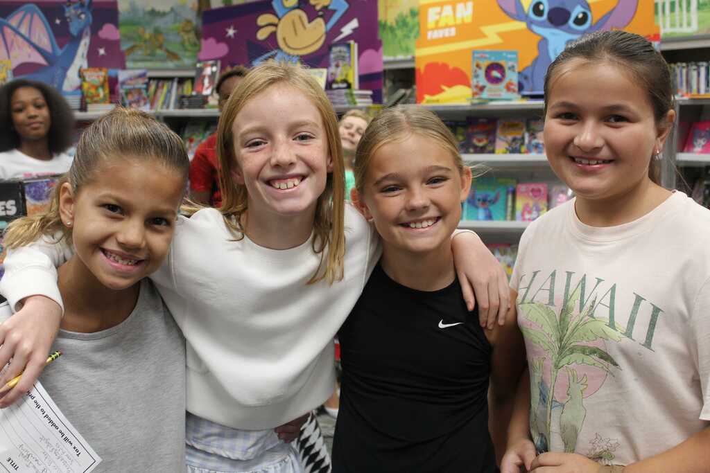 Four young girls smile and pose for a photo in a brightly lit bookstore. Shelves filled with colorful books are in the background.