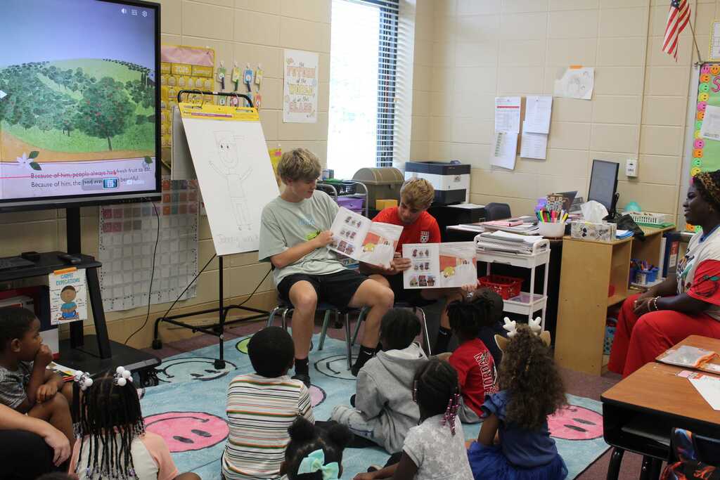 Children sit on a rug in a classroom, listening to a teacher read aloud while a monitor displays an animated scene.