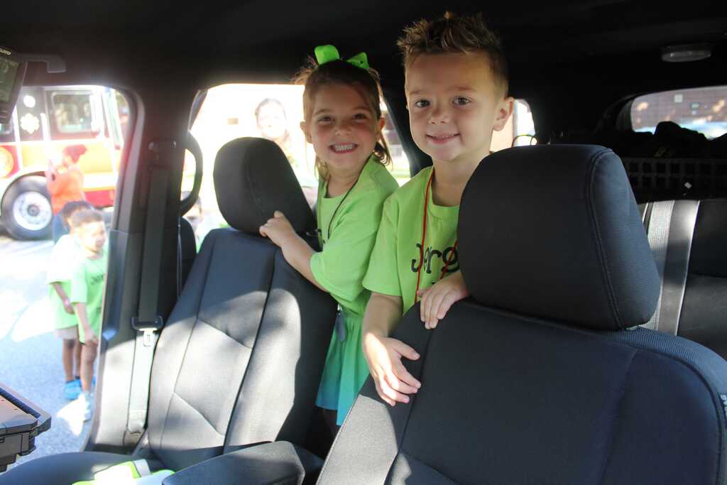Two children in green shirts sit in a vehicle's front seats, smiling. Behind them is a partially visible emergency vehicle.