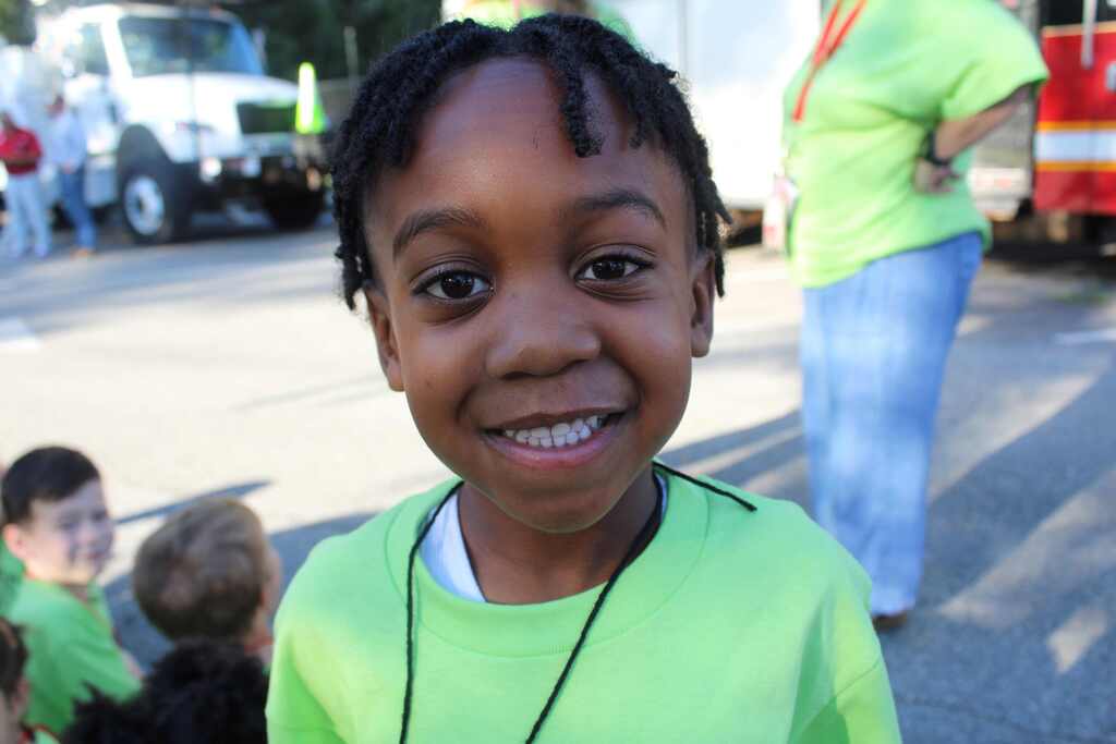 A young girl smiles with her teeth showing, wearing a green t-shirt. Behind her, a fire truck is visible.