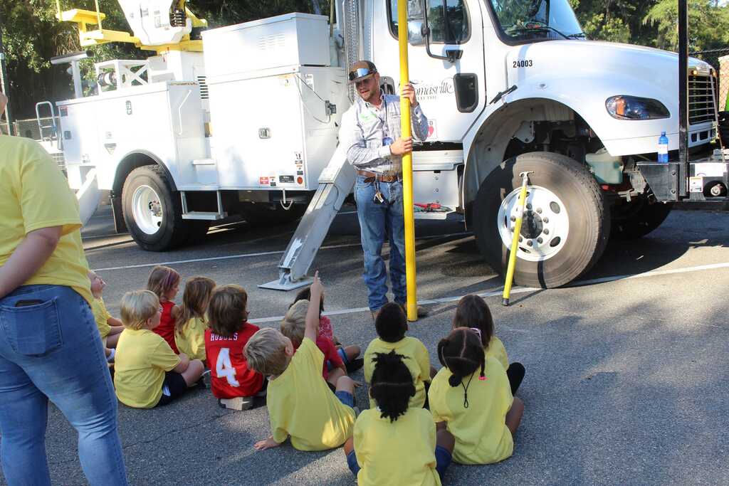 A group of children sits in a circle, with a utility worker standing beside a large vehicle.