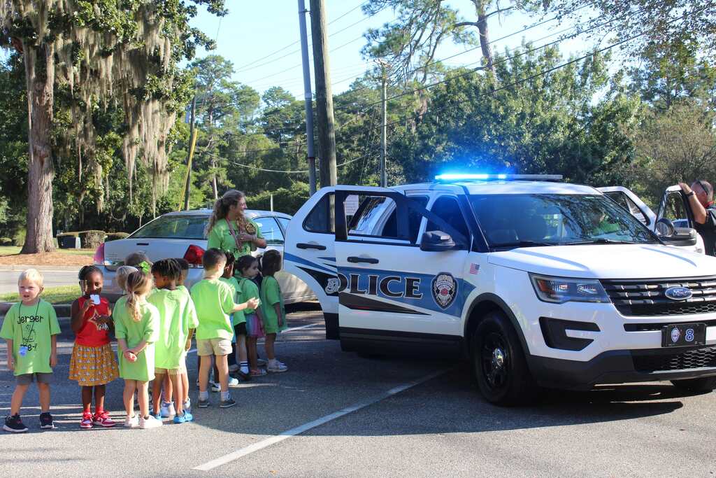 Children in green shirts gather around a police SUV with flashing lights. Tall trees in the background.