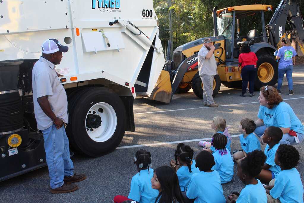 A man stands near a garbage truck as children in matching blue shirts sit in a semi-circle.