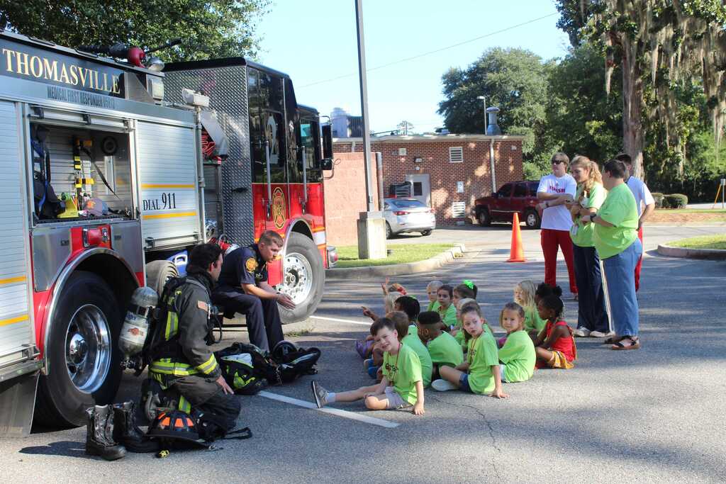 Children sit on the pavement near a fire truck. A firefighter is seated nearby, speaking with them.