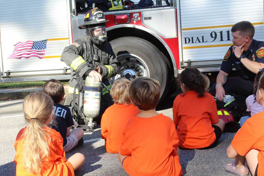 A group of children sits in front of a fire truck, with a firefighter speaking to them.