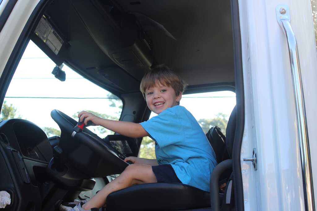 A child in a blue shirt sits in a truck, holding the steering wheel. The truck's door is open.
