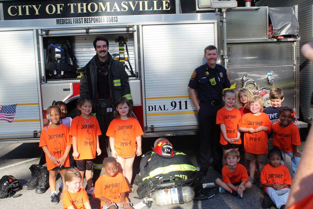 Firemen pose with a group of young children in orange T-shirts in front of a fire truck.