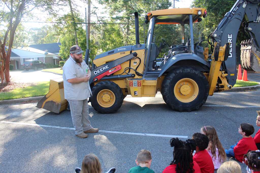 A man stands before a yellow backhoe in a parking lot with a crowd of children seated.