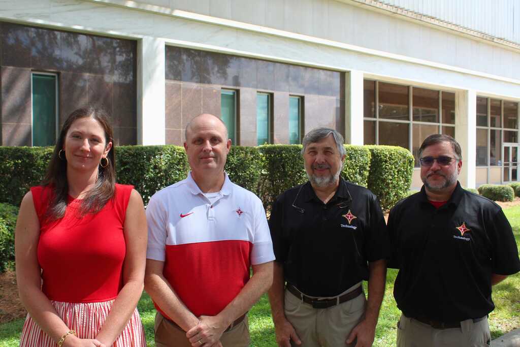 Four people stand in front of a building with glass windows, surrounded by bushes. Each wears a different-colored shirt.