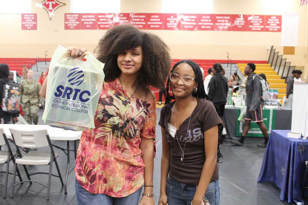 Two women stand in a crowded room with tables and people. One holds a bag with the letters SRTC.